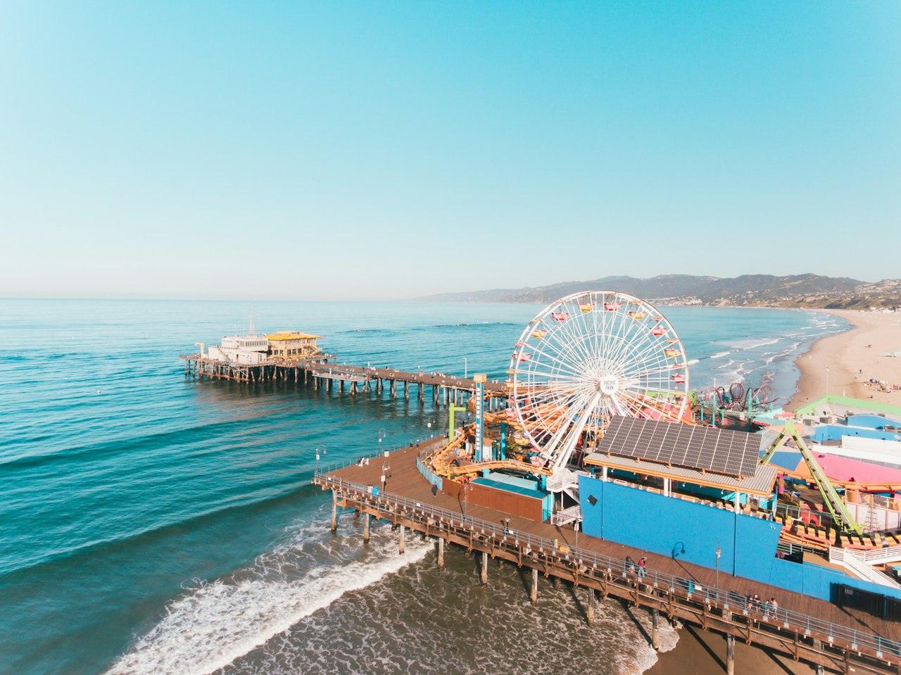 Aerial view of the Santa Monica Pier and its Ferris wheel over turquoise water, with the beach and Santa Monica Mountains beyond