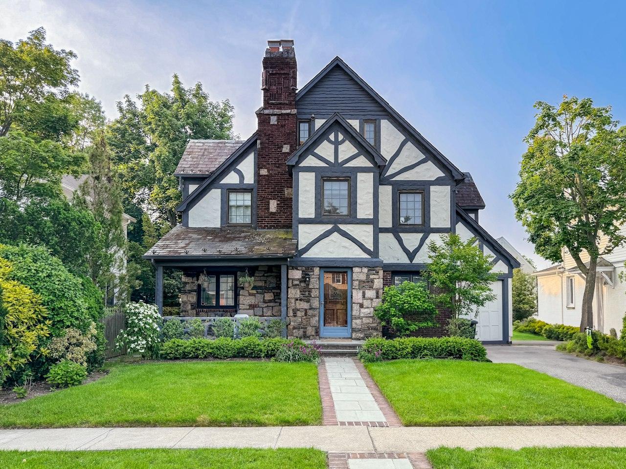A Tudor Revival house with steep gables, dark half-timbering over white stucco, a stone base and a tall brick chimney